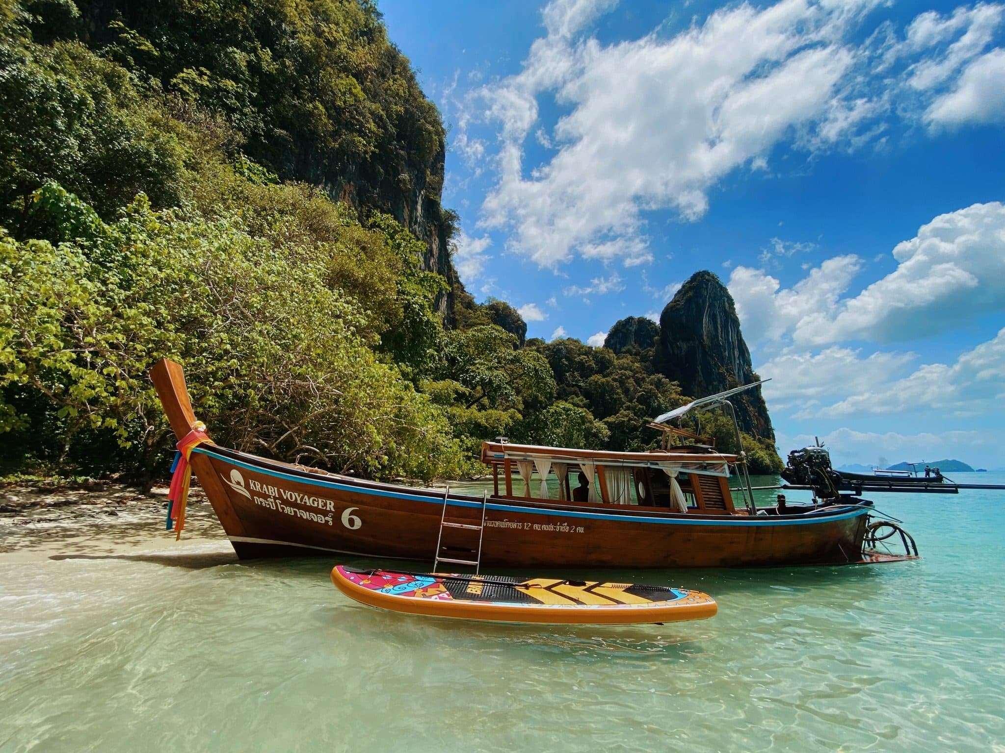 Krabi Voyager longtail boat moored at a quiet beach below limestone cliffs, with a paddleboard in the shallows
